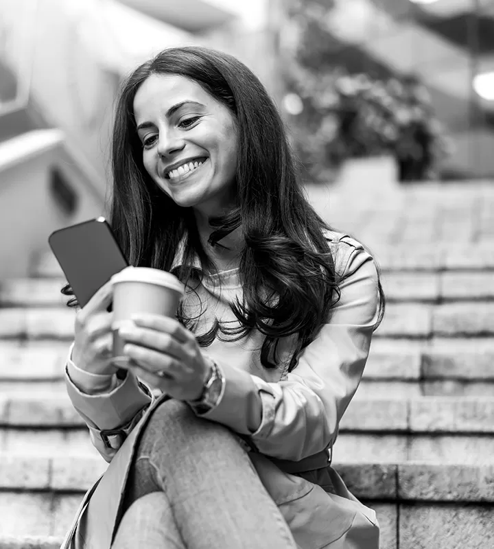 A happy woman looking at her phone while drinking a coffee